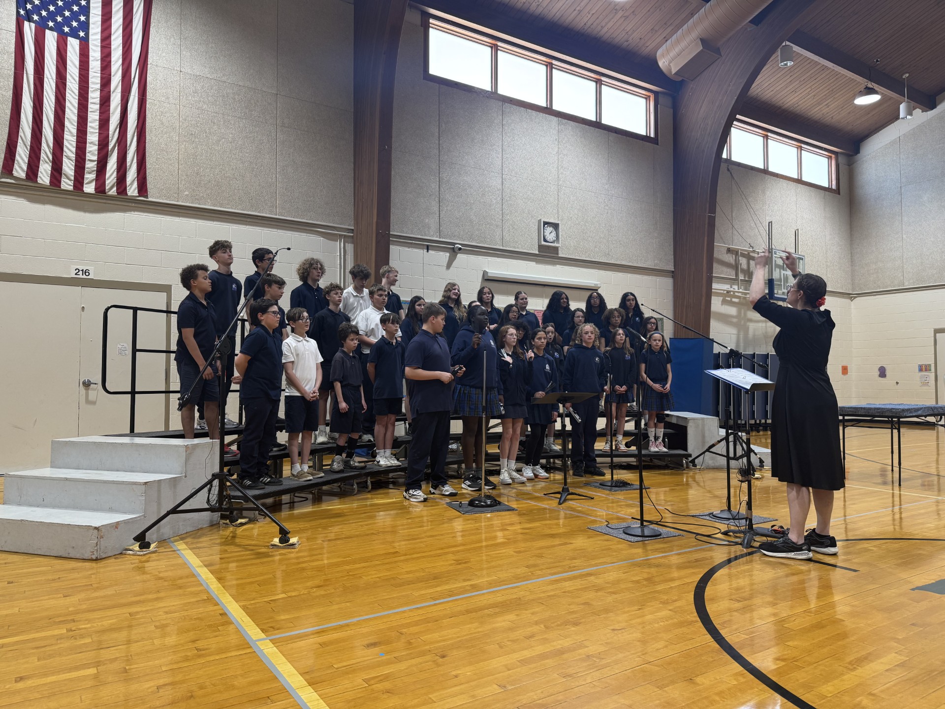 School choir of students in navy uniforms singing on risers in a gym, conductor directing at front with microphone stand nearby.